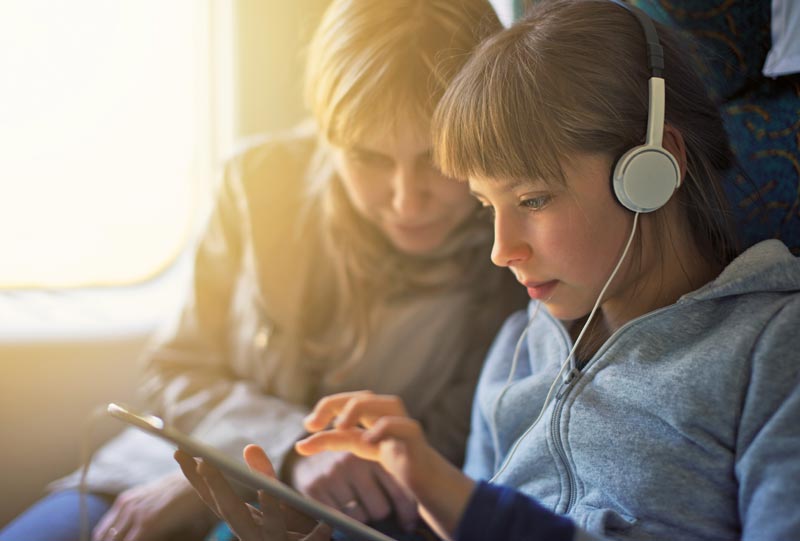 Νεαρή κοπέλα σε τρένο φοράει ακουστικά και ασχολείται με την ταμπλέτα της, ενώ δίπλα της κάθεται μια μεγαλύτερη γυναίκα.